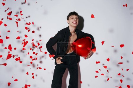 Foto de Hombre sonriente en jeans y camisa sosteniendo globo rojo cerca de confeti en forma de corazón sobre fondo gris - Imagen libre de derechos
