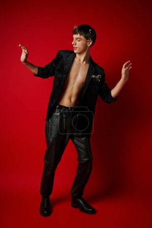 Photo for Full length of trendy young man in black attire with flowers in his hair gesturing on red background - Royalty Free Image