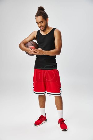 joyous appealing african american sportsman holding basketball and looking at phone, online betting