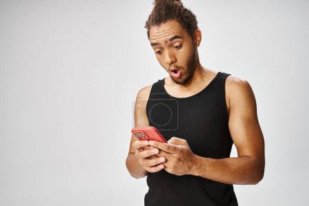 surprised athletic african american man looking at his smartphone on gray backdrop, online betting
