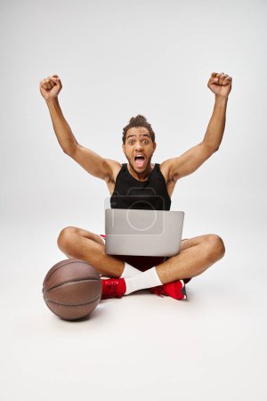 Téléchargez les photos : Homme afro-américain joyeux assis sur le sol avec un ordinateur portable acclamant en regardant la caméra, paris en ligne - en image libre de droit