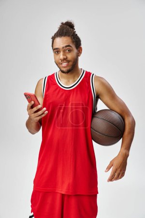 freudiger afrikanisch-amerikanischer Mann hält Basketball und Smartphone in der Hand und schaut in die Kamera, Online-Wetten