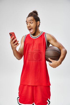 shocked african american man in sportwear holding basketball and looking at phone, online betting