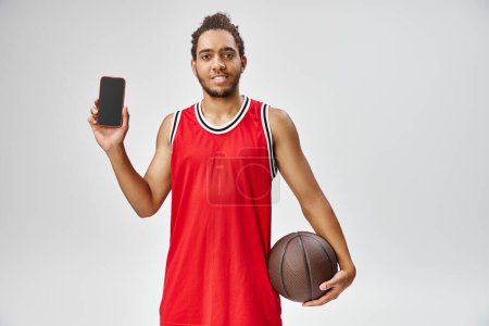 cheerful athletic african american man holding basketball and smiling at camera, online betting
