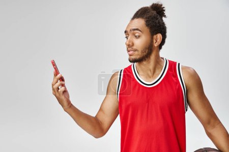 handsome jolly african american man holding basketball and looking at his phone, online betting