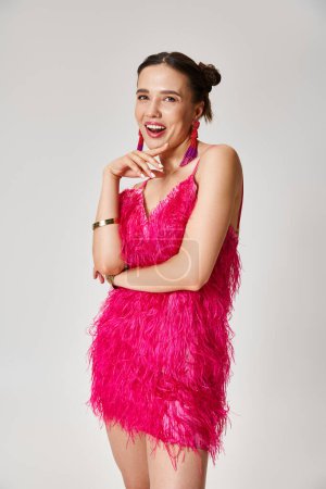 Téléchargez les photos : Jeune dame lumineuse en robe rose élégante posant joyeusement regardant à la caméra sur fond gris - en image libre de droit