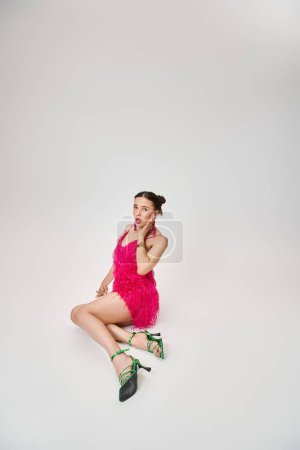 Chica juguetona en vestido rosa de moda y zapatos verdes tocando su mejilla y sentado sobre fondo gris