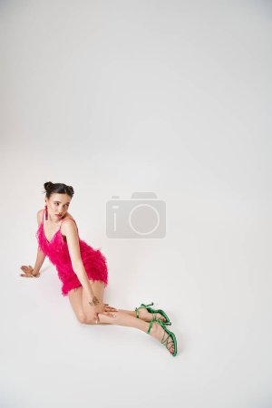 Pretty woman in pink dress touching her knee, sitting and looking away on grey background