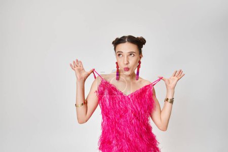 Happy woman in pink outfit looking away joyfully, holding straps of her dress on grey background