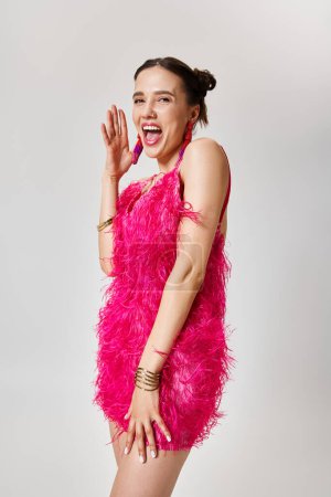 Mujer divertida en elegante vestido rosa se ríe, toca su mejilla, sobre fondo gris