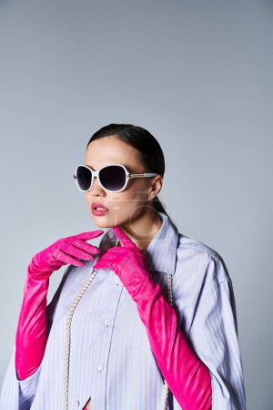 Schöne brünette Frau in trendiger Kleidung und Sonnenbrille, die im Studio ihr Kinn berührt