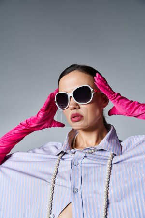 Téléchargez les photos : Brune fashion-forward en gants roses et lunettes de soleil tendance sur fond gris - en image libre de droit