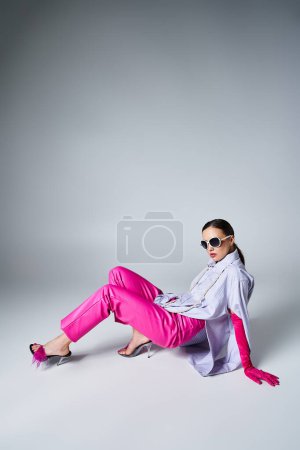 Foto de Mujer de moda con guantes rosas, pantalones de cuero y gafas de sol elegantes sobre fondo gris - Imagen libre de derechos