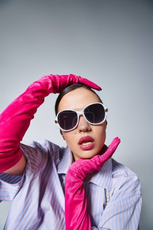 Femme à la mode avec des gants roses touchant élégamment son visage, portant des lunettes de soleil