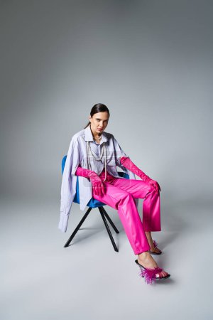 Foto de Chic chica morena en elegante traje rosa tocando sus piernas, sentado en la silla sobre fondo gris - Imagen libre de derechos