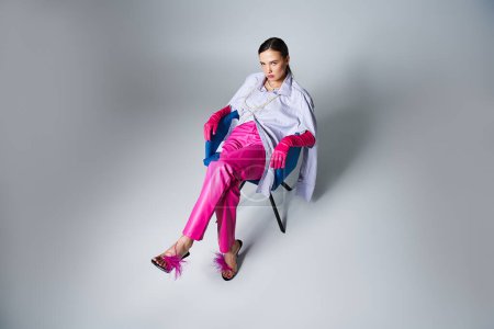 Foto de Chica morena con estilo en traje rosa de moda sentado en la silla sobre fondo gris, mirando a la cámara - Imagen libre de derechos