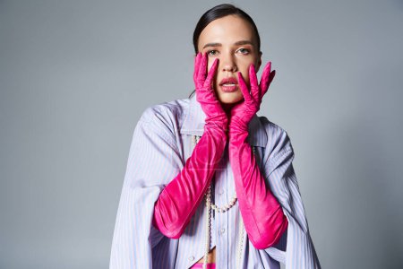 Portrait of fashion-forward brunette girl in pink gloves, touches her face on grey background