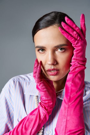 Pretty woman in stylish outfit and pink gloves touches her face while looking at camera on grey