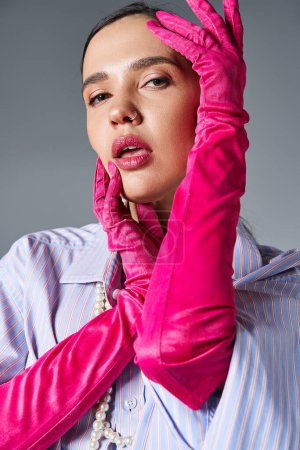 Brunette woman with piercing in stylish outfit and pink gloves, touches her face looking at camera