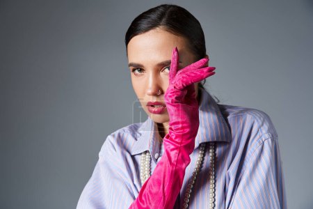 Fashionable woman with piercing in stylish outfit and pink gloves, covering her face with hand