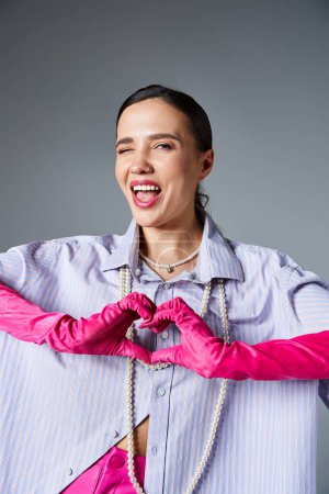 Téléchargez les photos : Souriant femme brune clignante avec des gants roses à la mode faisant coeur de la main isolé sur fond gris - en image libre de droit