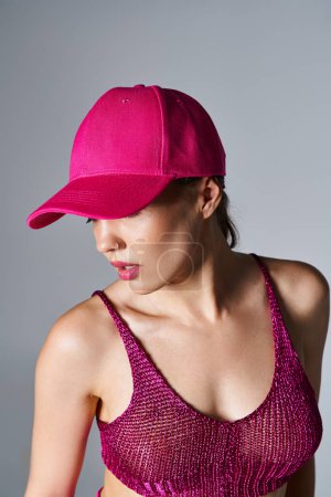 Brunette woman with piercing in stylish outfit with red top and pink cap, looking away on grey
