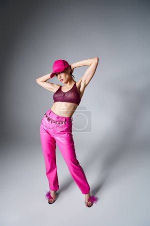 Full length photo of woman in red top, pink pants and cap on grey background with hands above head