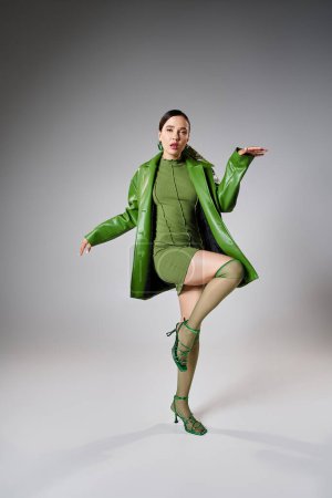Foto de Morena de moda en mini vestido verde, chaqueta de cuero, calcetines de rodilla bailando sobre fondo gris - Imagen libre de derechos