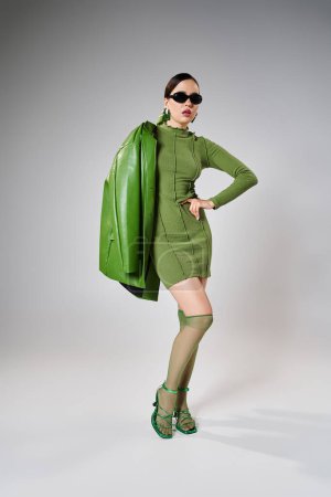 Brune à la mode en mini-robe verte, veste en cuir, chaussettes à genoux et chaussures, mains sur son corps