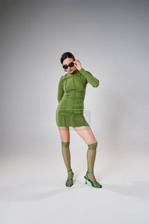 Foto de Mujer atractiva y elegante con traje verde y tocando sus gafas de sol de moda con una mano - Imagen libre de derechos