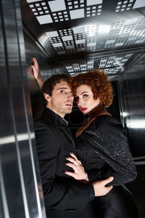 sexy schönes Paar in schickem schwarzen Kleid und Anzug, das sich im Fahrstuhl umarmt und in die Kamera schaut
