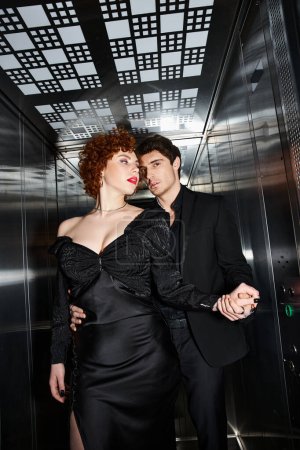 sexy appealing couple in chic black dress and suit hugging in elevator and looking at camera