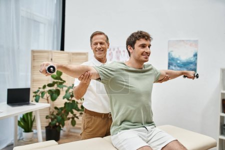 joyous mature doctor helping his cheerful handsome patient to use dumbbells during rehabilitation