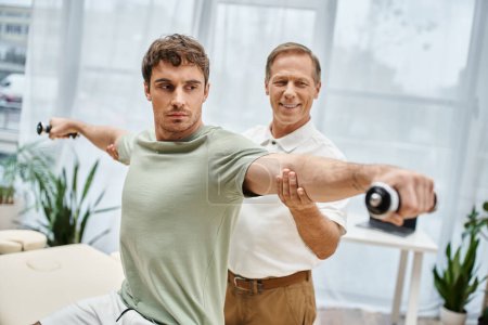 dedicated jolly mature doctor helping his patient with dumbbells on rehabilitation in hospital