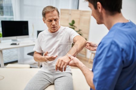 focus on mature patient in casual attire sitting next to his blurred doctor during rehabilitation