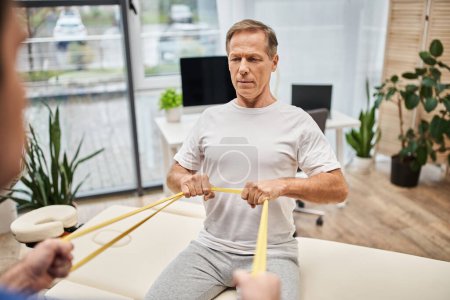 focus on handsome patient training with resistance band with his blurred doctor, rehabilitation