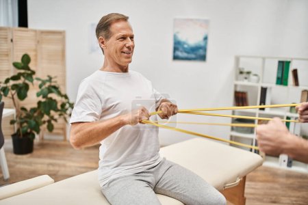 focus on mature jolly patient in casual attire training with resistance band with his blurred doctor