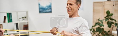 focus on mature jolly patient training with resistance band with his blurred doctor, banner