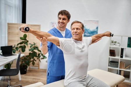 fröhlicher reifer Patient mit Kurzhanteln während der Reha mit Hilfe seines gutaussehenden lustigen Arztes