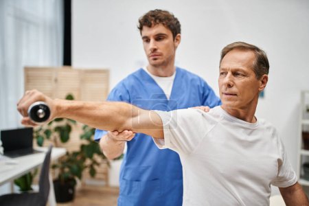 dedicated doctor in blue robe helping mature handsome patient with dumbbell during rehabilitation