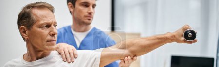 doctor devoto en bata azul ayudando a madurar paciente atractivo con mancuerna durante la rehabilitación
