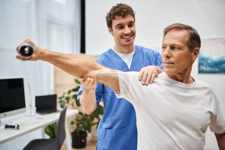 joyful doctor in blue robe helping his mature handsome patient to use dumbbell during rehabilitation