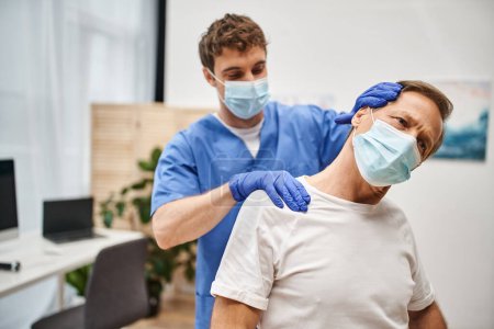 Téléchargez les photos : Médecin attrayant avec masque médical et gants aidant son patient à étirer pendant le rendez-vous - en image libre de droit