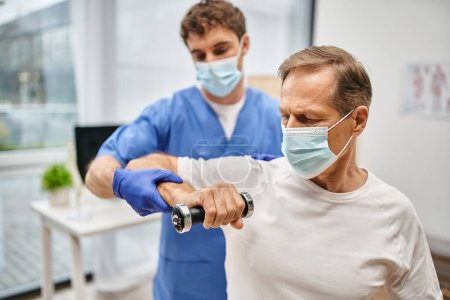 Téléchargez les photos : Dévoué beau médecin avec masque et gants aidant son patient à utiliser des haltères sur rendez-vous - en image libre de droit