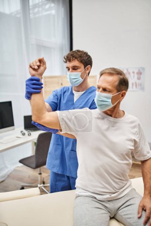 Fleißiger Arzt mit Maske und Handschuhen hilft erwachsenen Patienten, seine Muskeln auf der Station zu rehabilitieren
