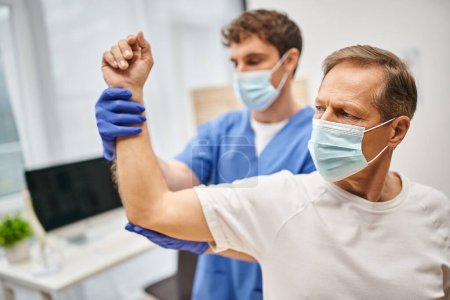 médico devoto con máscara médica y guantes que ayudan al paciente maduro a rehabilitar su cuerpo en la sala