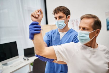 médico trabajador en guantes médicos y máscara rehabilitando a su paciente maduro en el hospital