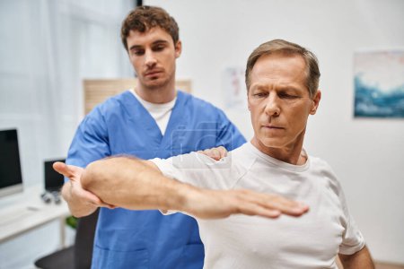 bien parecido doctor dedicado en bata azul ayudando a su paciente a rehabilitar su cuerpo en la sala