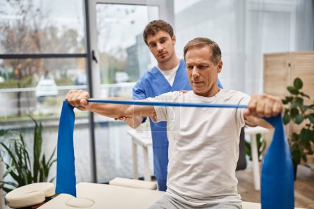 hardworking doctor in blue uniform helping his mature patient with resistance band on rehabilitation