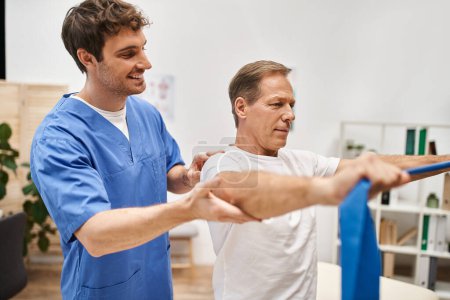 cheerful handsome doctor in blue robe helping his mature patient to use resistance band in hospital